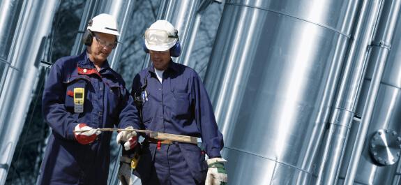 Két dolgozó kék munkaruhában áll egy gázvezeték előtt